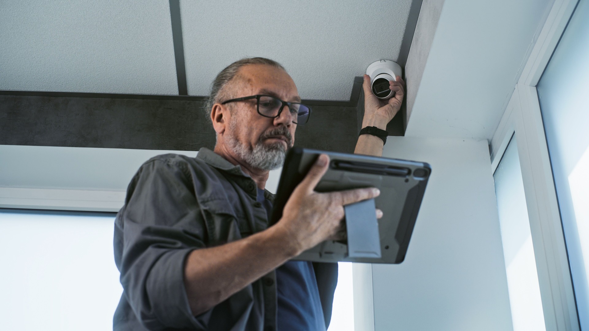 Male mature installer sets up CCTV camera in office room using tablet computer